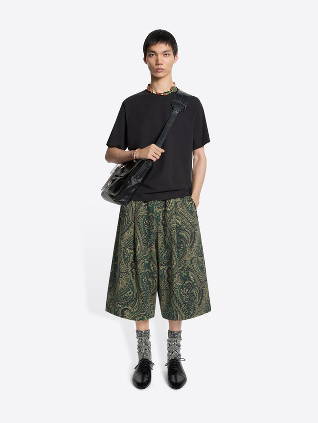 Person wearing a black t-shirt, green patterned shorts, and carrying a black bag on a white background