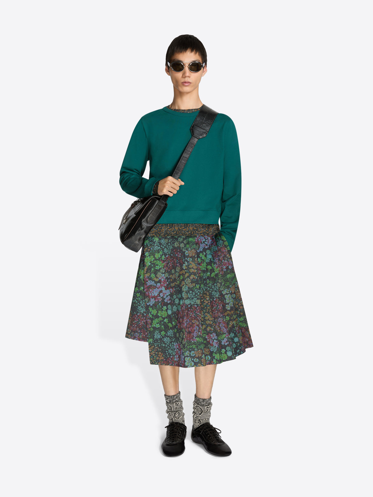 Person wearing a teal sweater and patterned skirt with a black bag on a white background