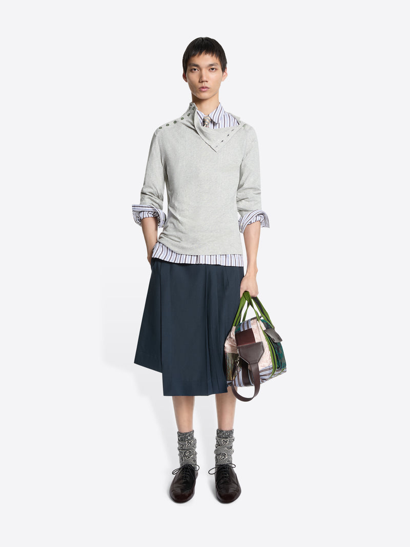 Person wearing a light gray sweater with rolled-up sleeves, a striped shirt, dark skirt, and holding a multicolored bag on a white background.