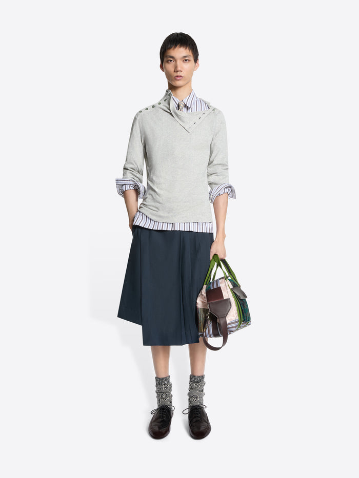 Person wearing a light gray sweater with rolled-up sleeves, a striped shirt, dark skirt, and holding a multicolored bag on a white background.