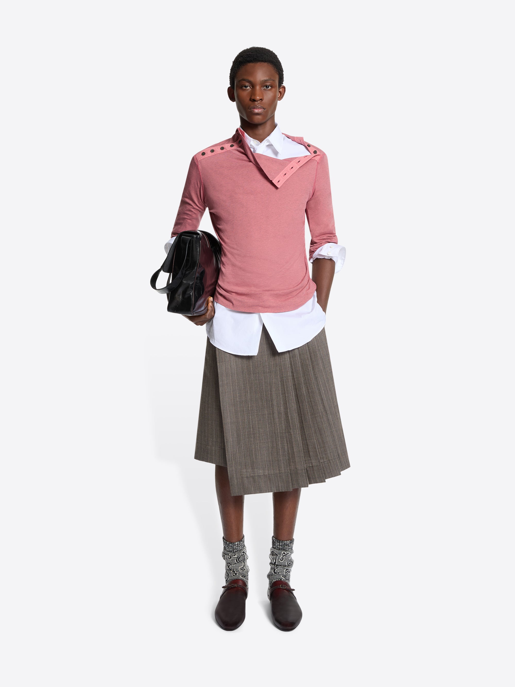 Person wearing a pink turtleneck tee, white shirt, brown pleated skirt, and brown shoes holding a black bag on a white background