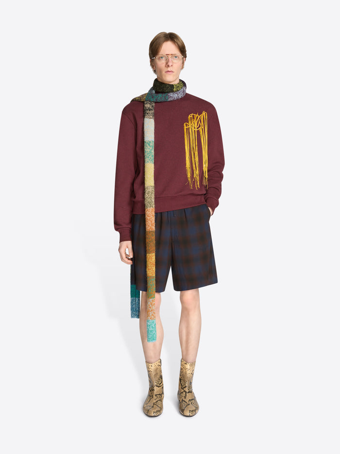 A model wearing a burgundy sweatshirt with yellow fringe embroidery, dark plaid shorts, a long multicolored knitted scarf, and tan snakeskin-pattern ankle boots against a light gray background.
