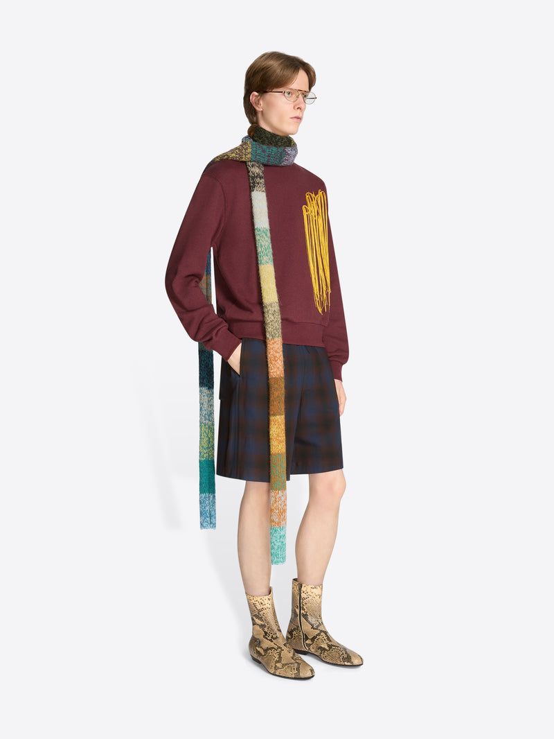 A model wearing a burgundy sweatshirt with yellow fringe embroidery, dark plaid shorts, a long multicolored knitted scarf, and tan snakeskin-pattern ankle boots against a light gray background.