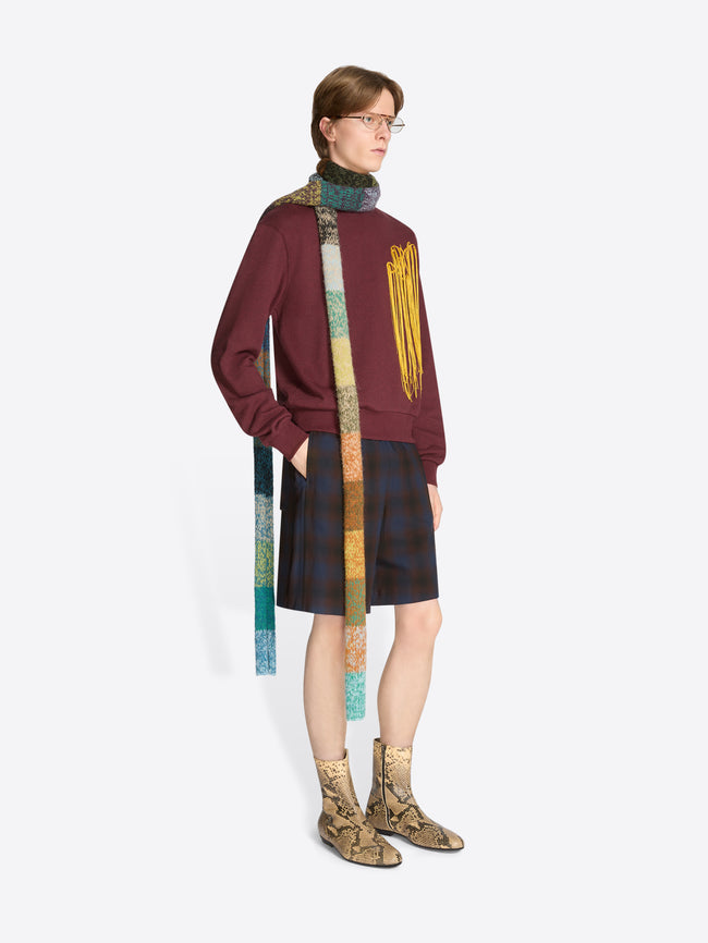 A model wearing a burgundy sweatshirt with yellow fringe embroidery, dark plaid shorts, a long multicolored knitted scarf, and tan snakeskin-pattern ankle boots against a light gray background.