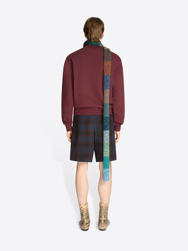 Back view of a model wearing a burgundy sweatshirt with yellow fringe embroidery, dark plaid shorts, a long multicolored knitted scarf, and tan snakeskin-pattern ankle boots against a light gray background.
