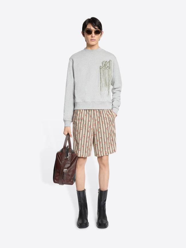Model wearing a light gray sweatshirt with dangling embroidered details, patterned knee-length shorts, and black mid-calf boots, holding a brown leather bag in one hand and stands against a light gray background.