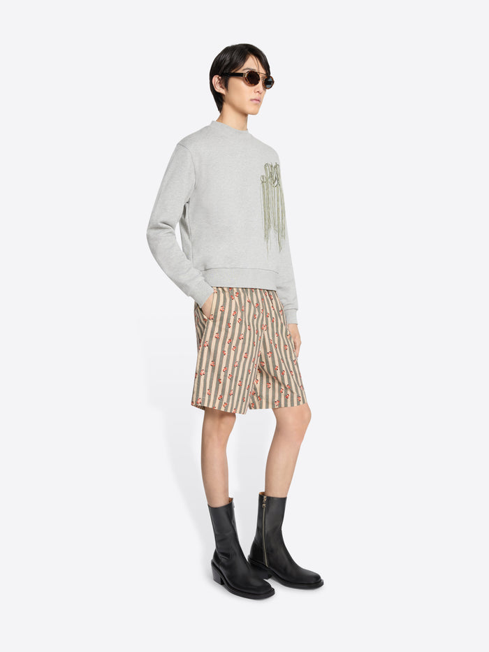 Model wearing a light gray sweatshirt with dangling embroidered details, patterned knee-length shorts, and black mid-calf boots, holding a brown leather bag in one hand and stands against a light gray background.