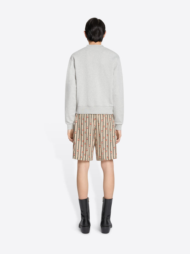 Back view of a model wearing a light gray sweatshirt with dangling embroidered details, patterned knee-length shorts, and black mid-calf boots, holding a brown leather bag in one hand and stands against a light gray background.
