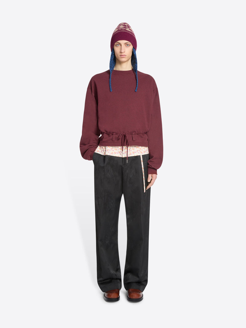 A model stands against a light gray background wearing a burgundy cropped sweatshirt with a drawstring waist, black wide-leg pants, brown shoes, and a burgundy hat with blue ear flaps.