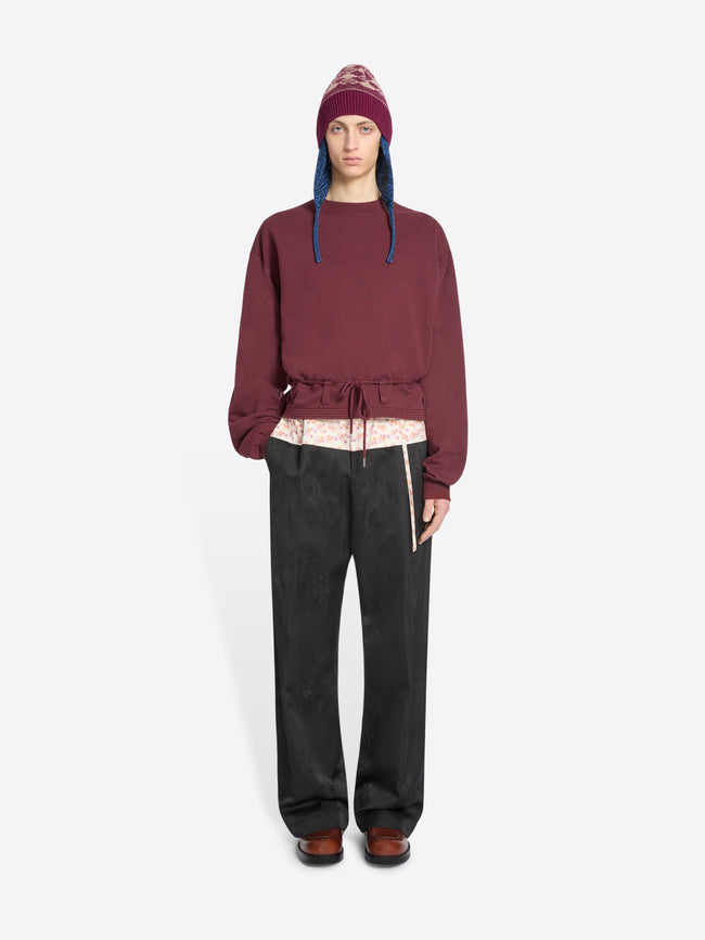 A model stands against a light gray background wearing a burgundy cropped sweatshirt with a drawstring waist, black wide-leg pants, brown shoes, and a burgundy hat with blue ear flaps.