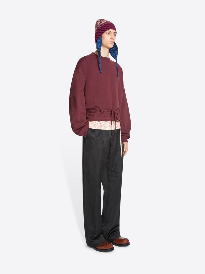 A model stands against a light gray background wearing a burgundy cropped sweatshirt with a drawstring waist, black wide-leg pants, brown shoes, and a burgundy hat with blue ear flaps.