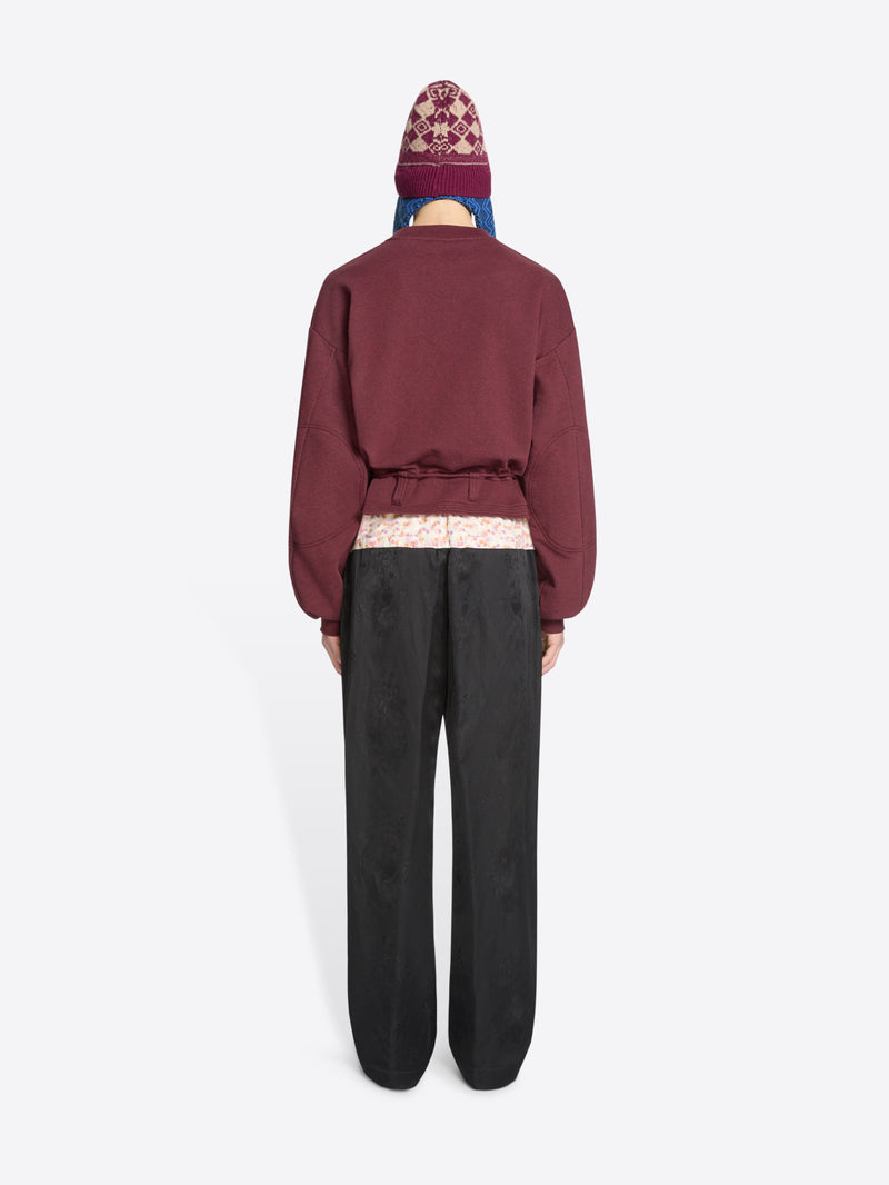 Back view of a model standing against a light gray background wearing a burgundy cropped sweatshirt with a drawstring waist, black wide-leg pants, brown shoes, and a burgundy hat with blue ear flaps.