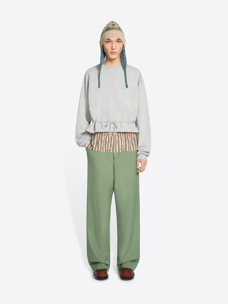 A model stands against a light gray background wearing a light gray long-sleeve sweatshirt with a ruffled hem, sage green wide-leg pants, brown shoes, and a knit hat.
