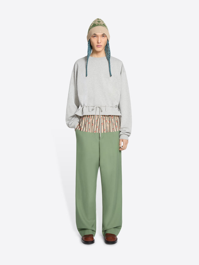 A model stands against a light gray background wearing a light gray long-sleeve sweatshirt with a ruffled hem, sage green wide-leg pants, brown shoes, and a knit hat.