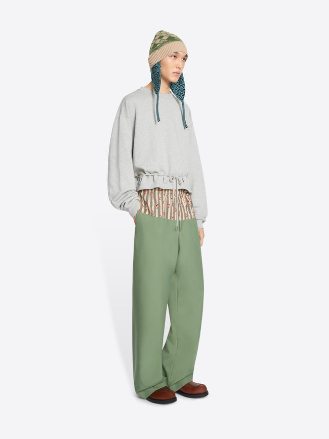 A model stands against a light gray background wearing a light gray long-sleeve sweatshirt with a ruffled hem, sage green wide-leg pants, brown shoes, and a knit hat.