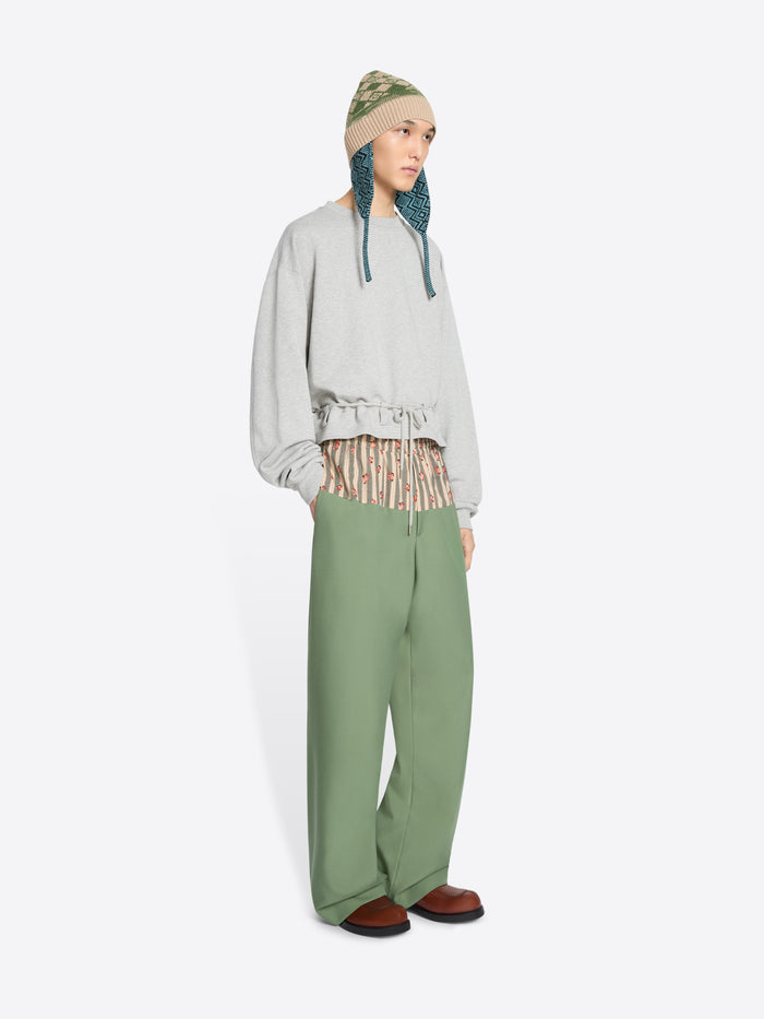 A model stands against a light gray background wearing a light gray long-sleeve sweatshirt with a ruffled hem, sage green wide-leg pants, brown shoes, and a knit hat.