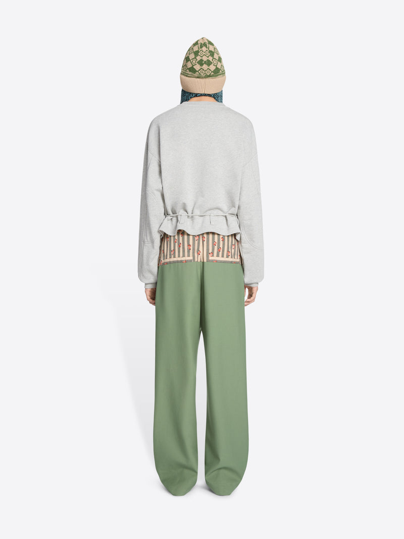Back view of a model standing against a light gray background wearing a light gray long-sleeve sweatshirt with a ruffled hem, sage green wide-leg pants, brown shoes, and a knit hat.