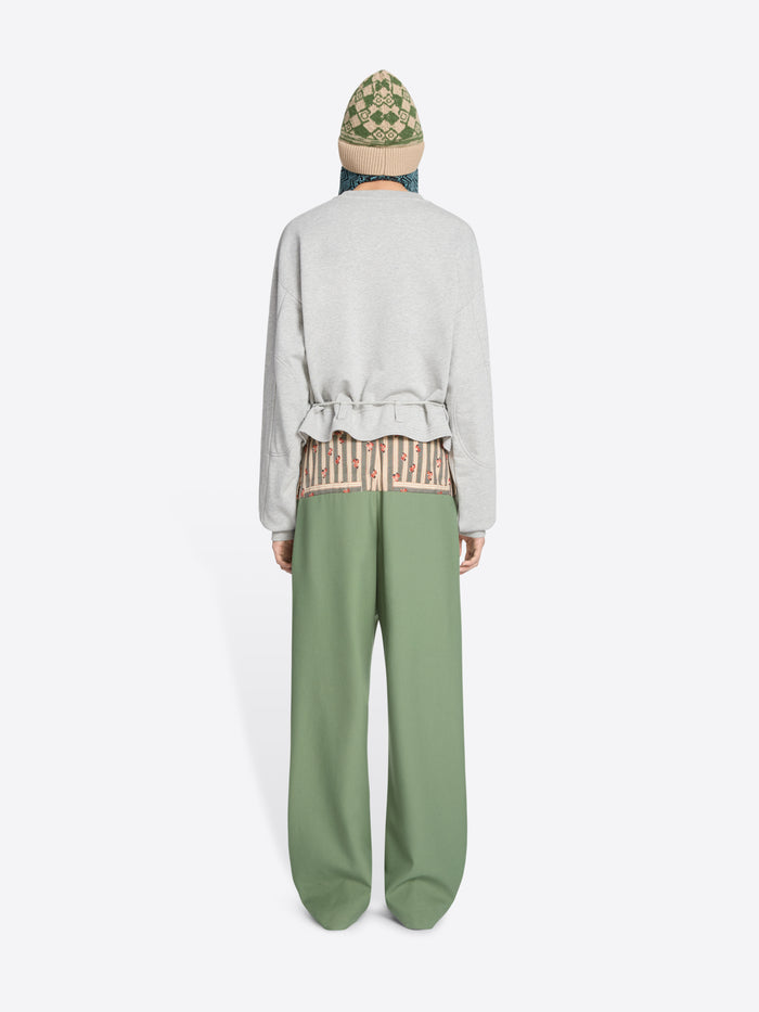Back view of a model standing against a light gray background wearing a light gray long-sleeve sweatshirt with a ruffled hem, sage green wide-leg pants, brown shoes, and a knit hat.