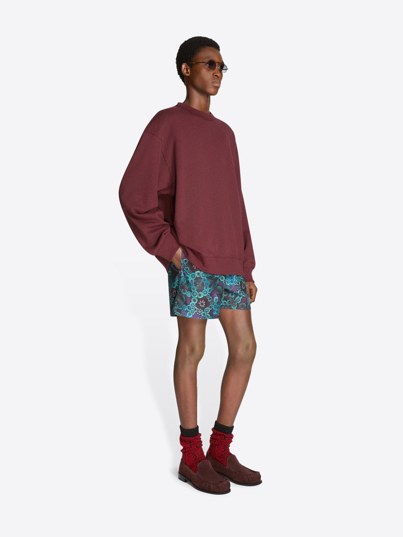 Person wearing a burgundy sweatshirt, patterned shorts, and brown shoes on a white background