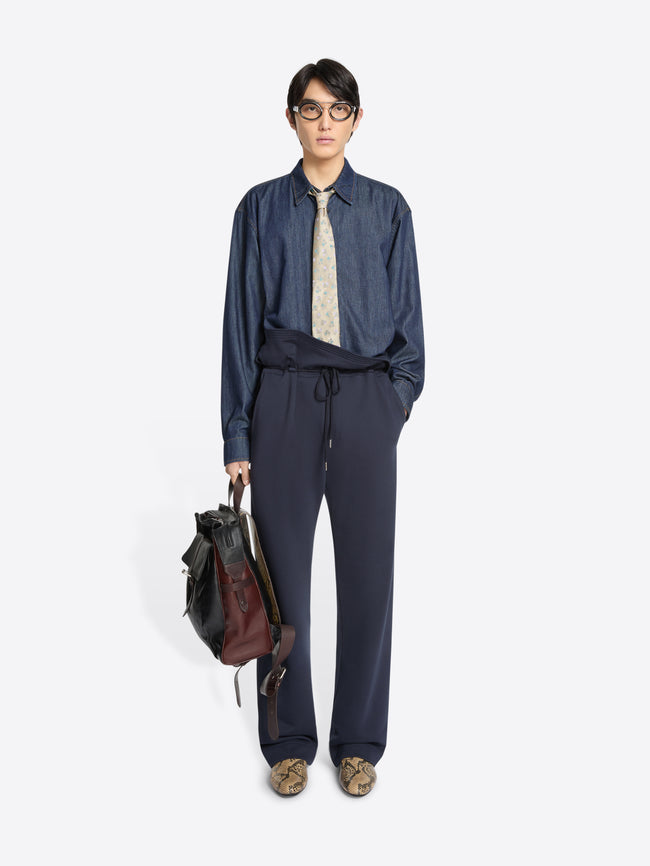 Person wearing a blue denim shirt and navy blue pants holding a leather backpack on a white background