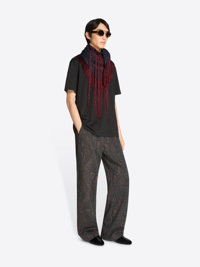 Person wearing a black t-shirt with a long navy scarf with red fringe, patterned pants, and sunglasses on a white background