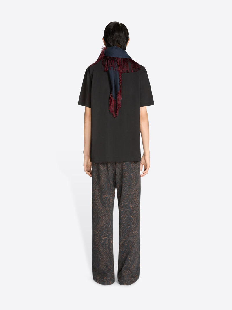 Back view of a person wearing a black t-shirt with a long navy scarf with red fringe, patterned pants, and sunglasses on a white background