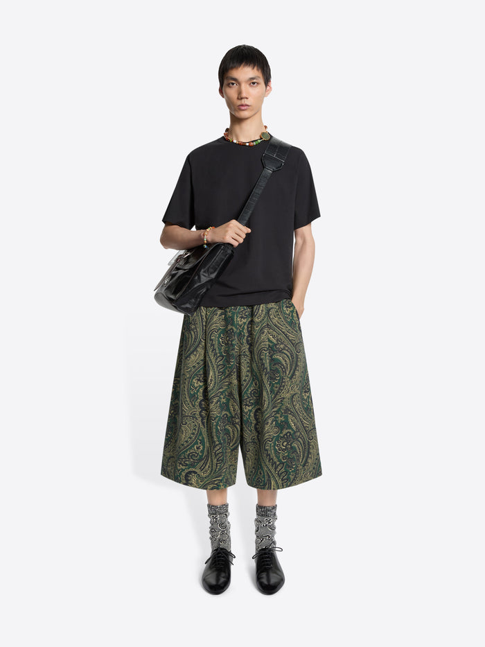 Person wearing a black t-shirt, green patterned shorts, and carrying a black bag on a white background