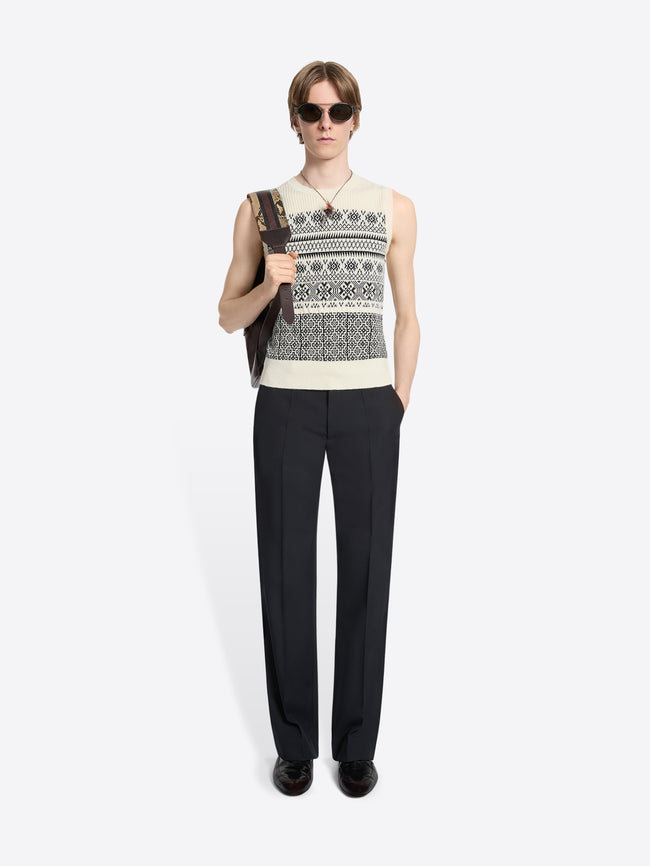 Person wearing a sleeveless patterned knitted top and black pants holding bag on a white background