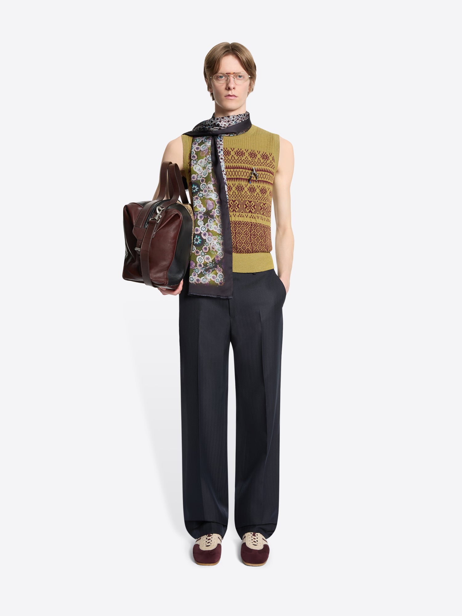Person wearing a sleeveless patterned top, black pants, patterned scarf and holding a brown bag on a white background