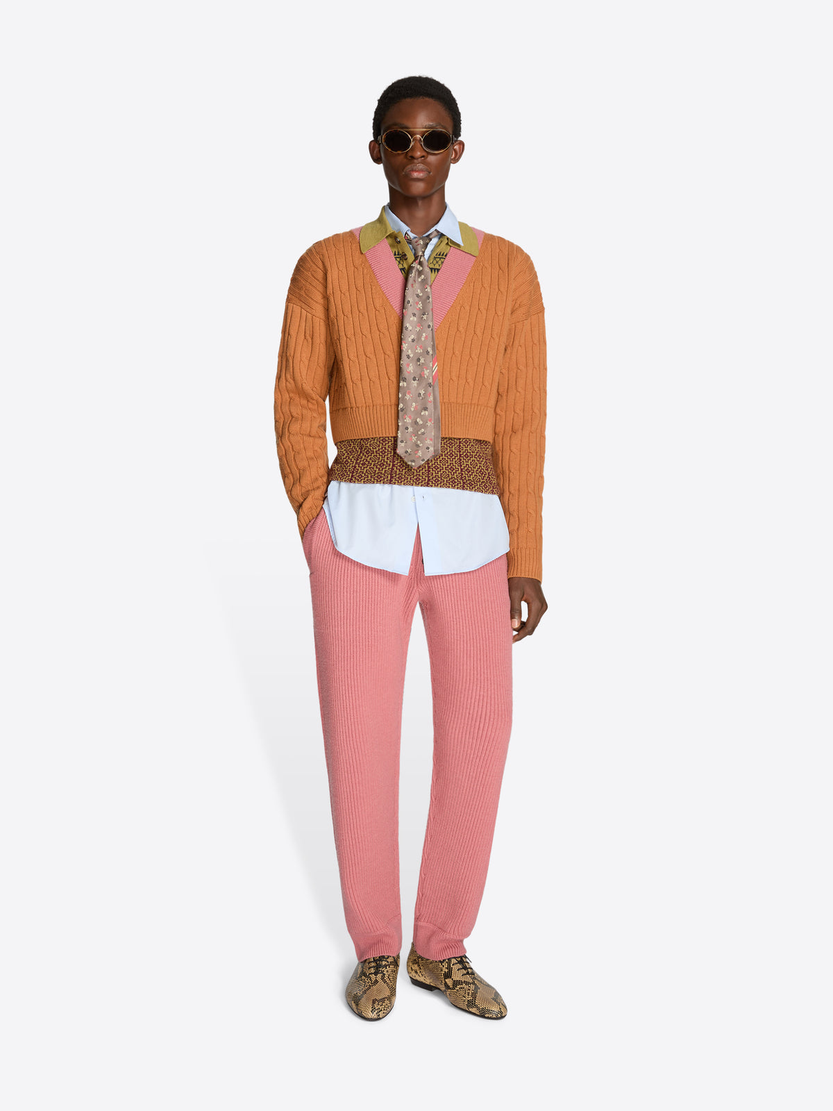 Person wearing an orange sweater with patterned band and collar, pink pants, and patterned shoes on a white background
