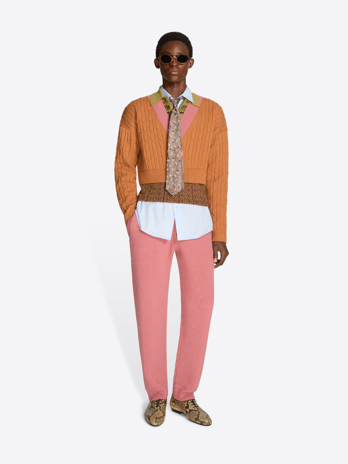 Person wearing an orange sweater with patterned band and collar, pink pants, and patterned shoes on a white background