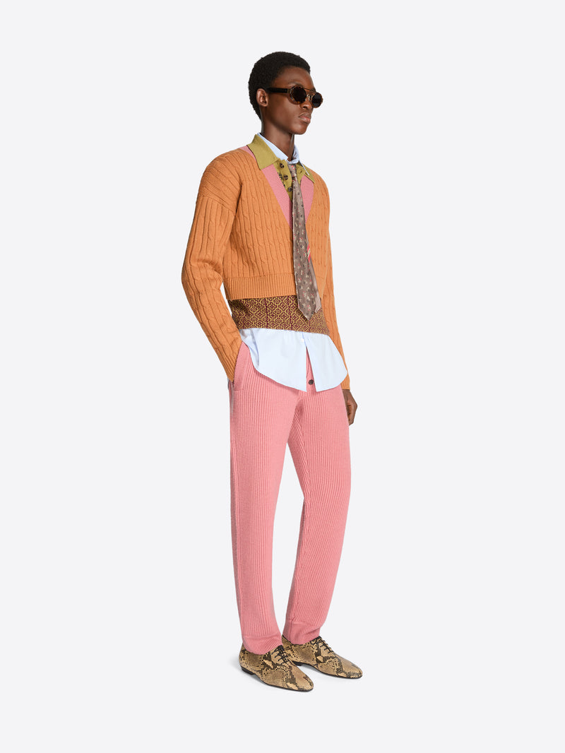 Person wearing an orange sweater with patterned band and collar, pink pants, and patterned shoes on a white background