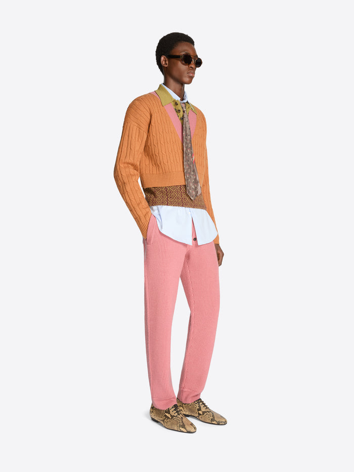 Person wearing an orange sweater with patterned band and collar, pink pants, and patterned shoes on a white background