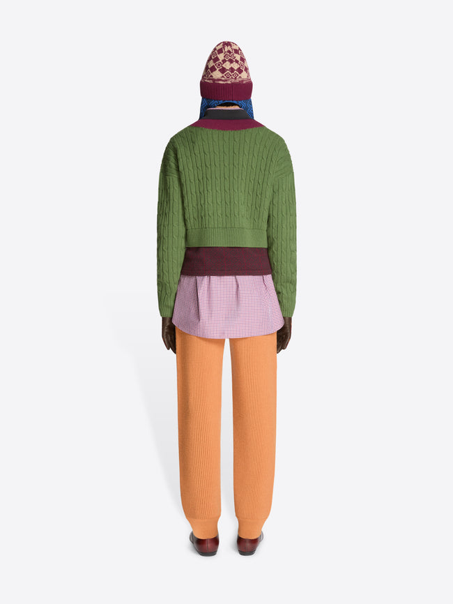 Back view of a person wearing a green sweater with patterned band and collar , purple checkered shirt, orange pants, and brown shoes on a white background
