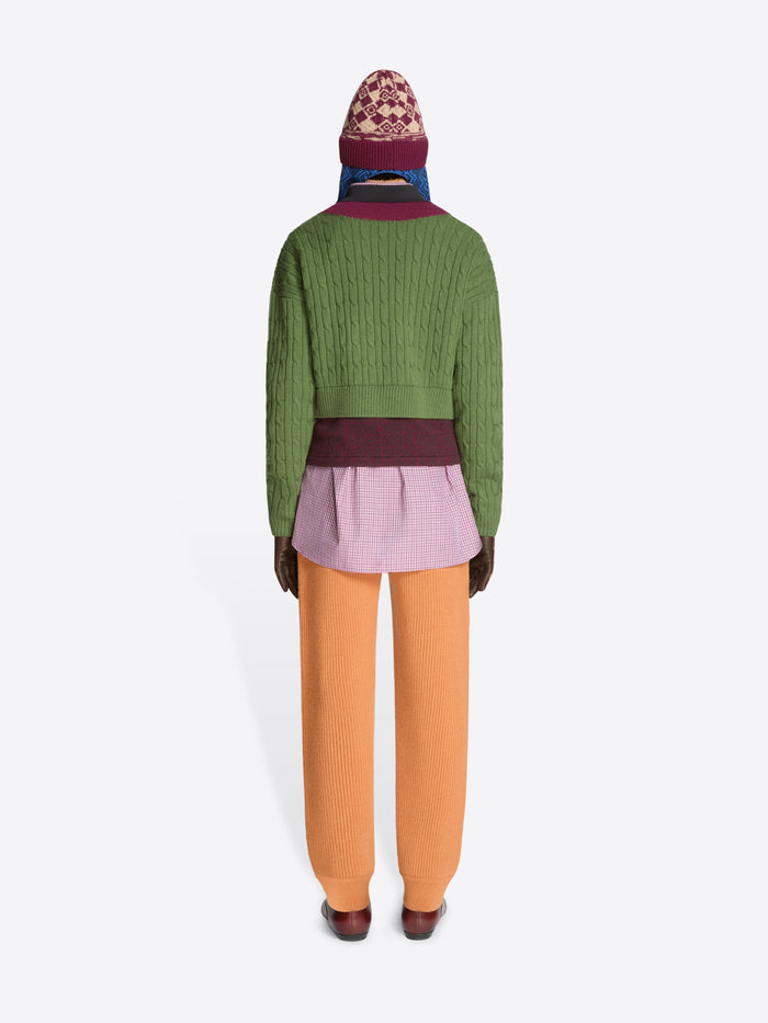Back view of a person wearing a green sweater with patterned band and collar , purple checkered shirt, orange pants, and brown shoes on a white background
