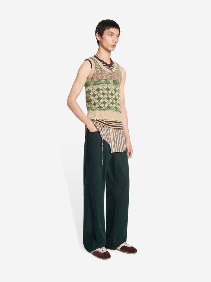Person wearing a patterned sleeveless top and dark pants on a white background