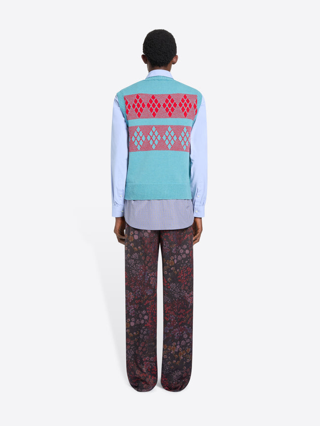 Back view of a person wearing a patterned knitted top over a blue shirt with a tie, patterned pants on a white background
