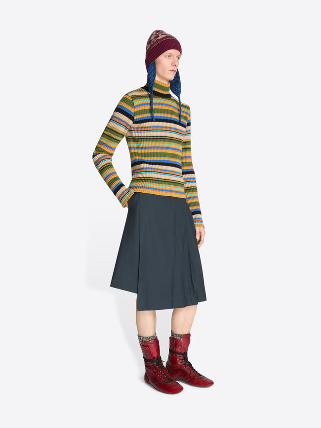 Person wearing a colorful striped sweater, dark skirt, patterned hat and red boots on a white background