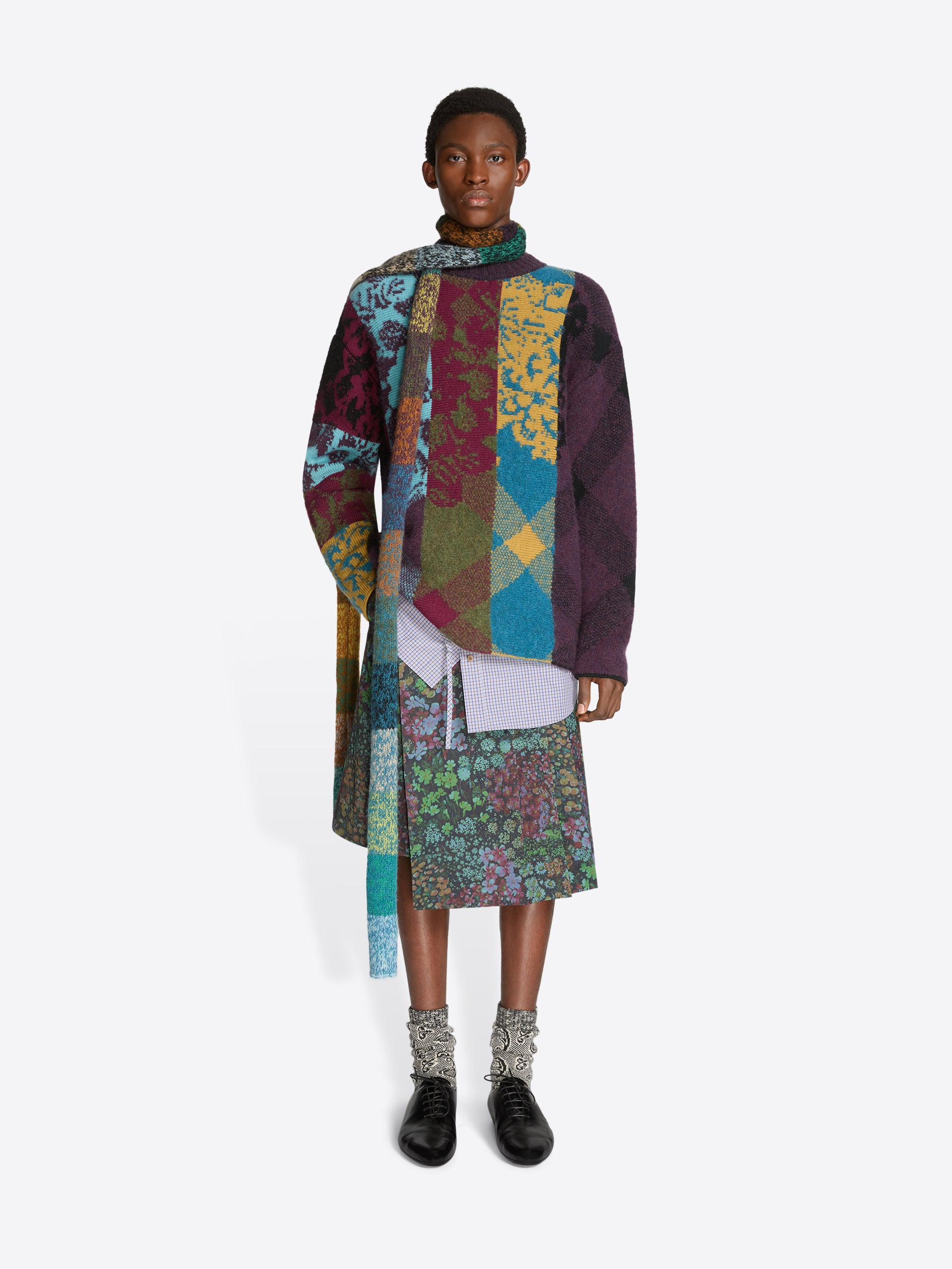 Person wearing a colorful patchwork sweater and floral patterned skirt on a white background