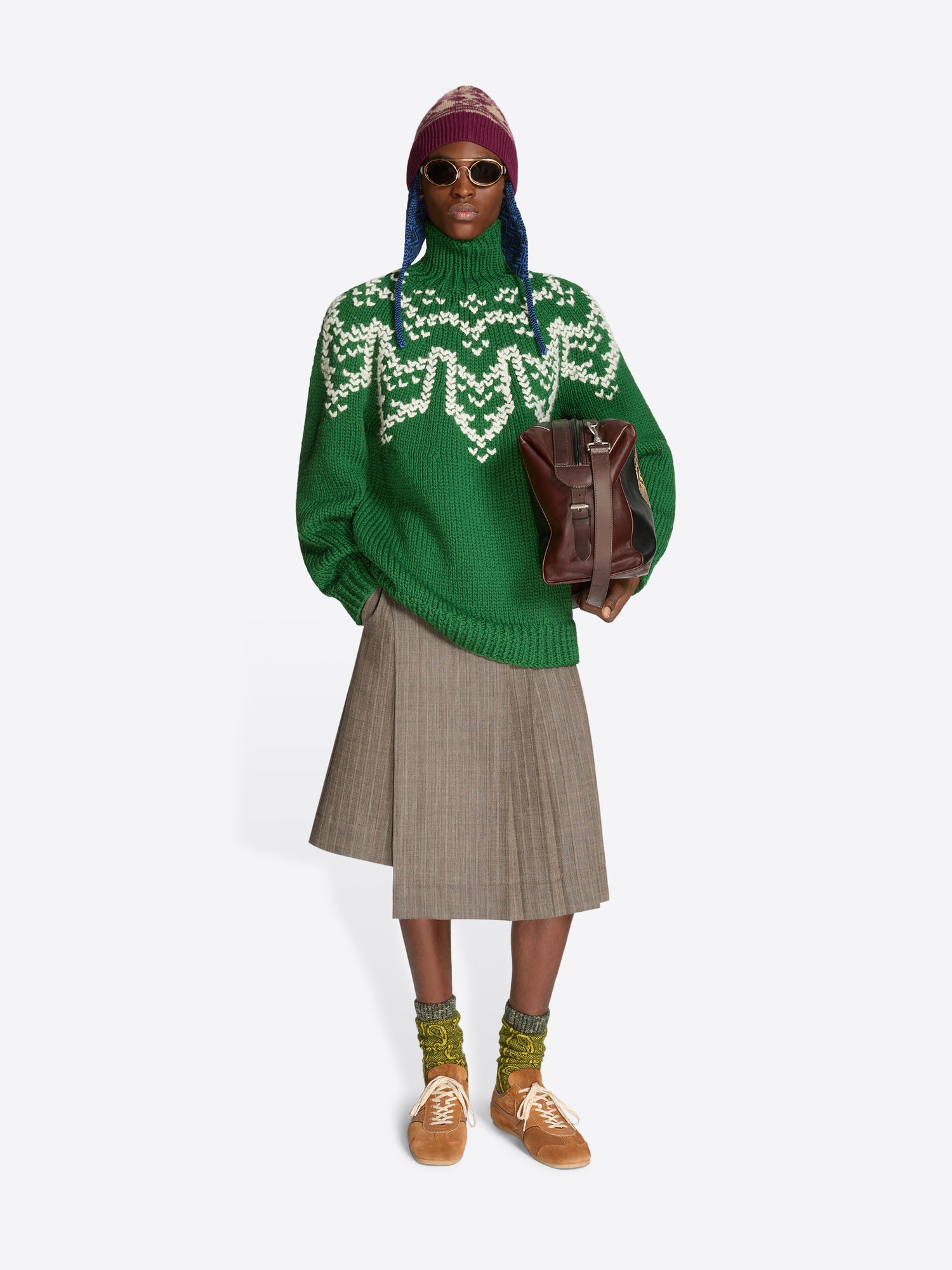 Model wearing a green patterned turtleneck sweater, gray pleated shorts, a knitted hat, sunglasses and a brown leather bag.
