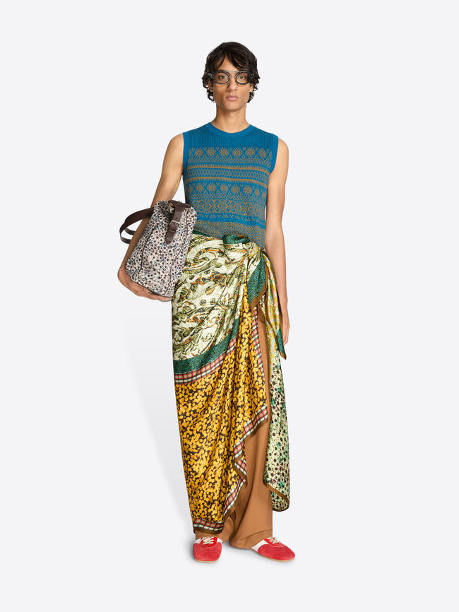 Person wearing a blue sleeveless top and patterned pareo with a handbag on a white background