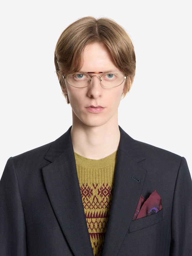 Person wearing a dark suit with a patterned sweater, silk pocket square and glasses on a white background