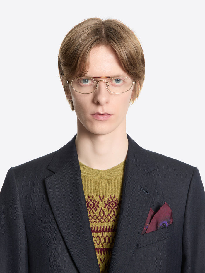 Person wearing a dark suit with a patterned sweater, silk pocket square and glasses on a white background