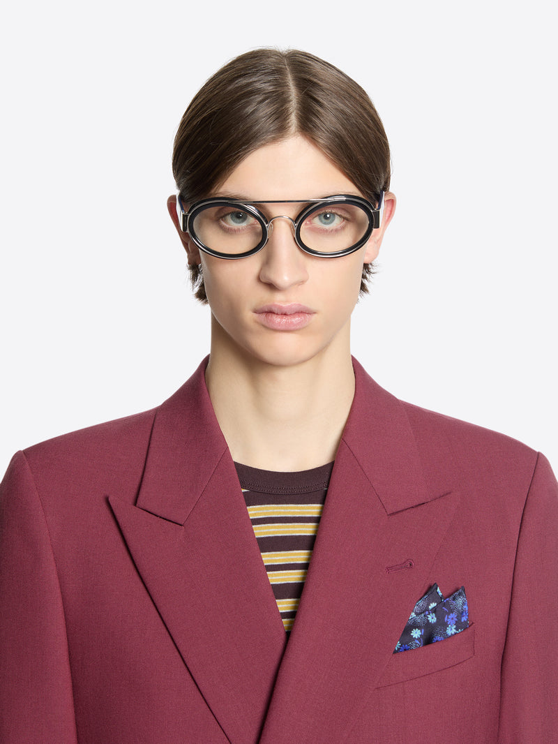 Person wearing a burgundy blazer with a striped shirt underneath, silk pocket square and glasses on a white background