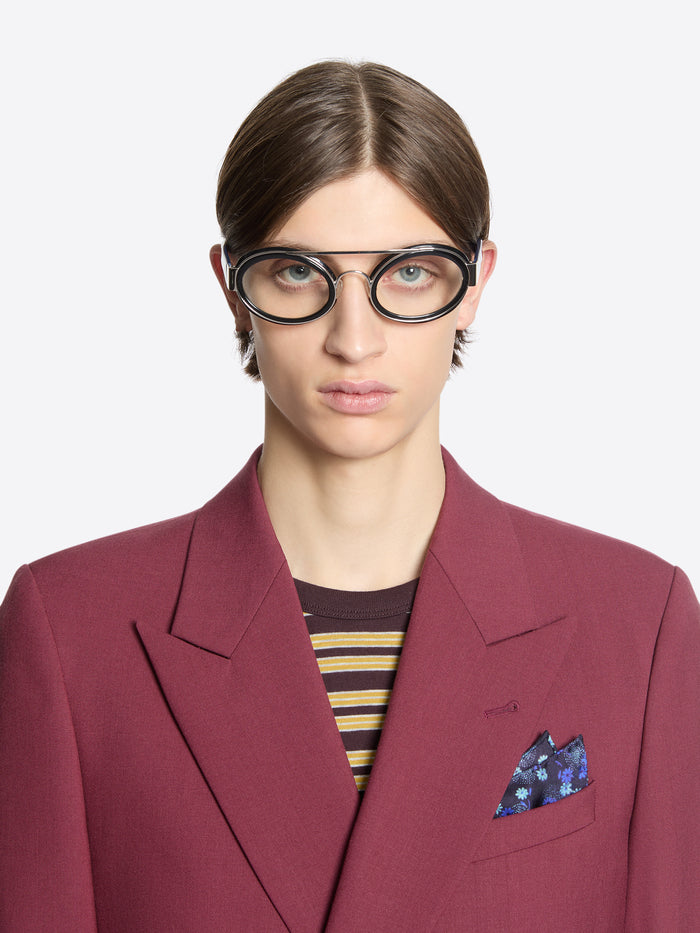 Person wearing a burgundy blazer with a striped shirt underneath, silk pocket square and glasses on a white background
