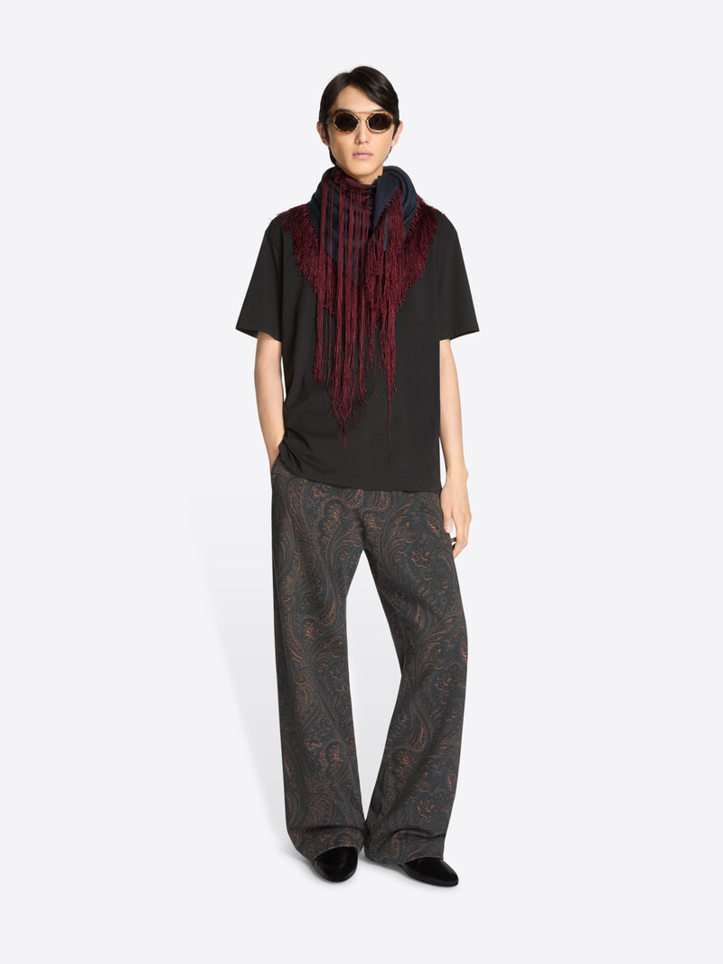 Person wearing a black t-shirt with a long scarf with red fringe, dark pants, and sunglasses on a white background