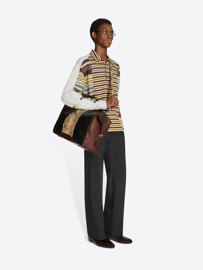 A model wearing a striped knit top with a white sleeve, dark wide-leg trousers, and carrying a large brown leather shoulder bag against a light gray background.