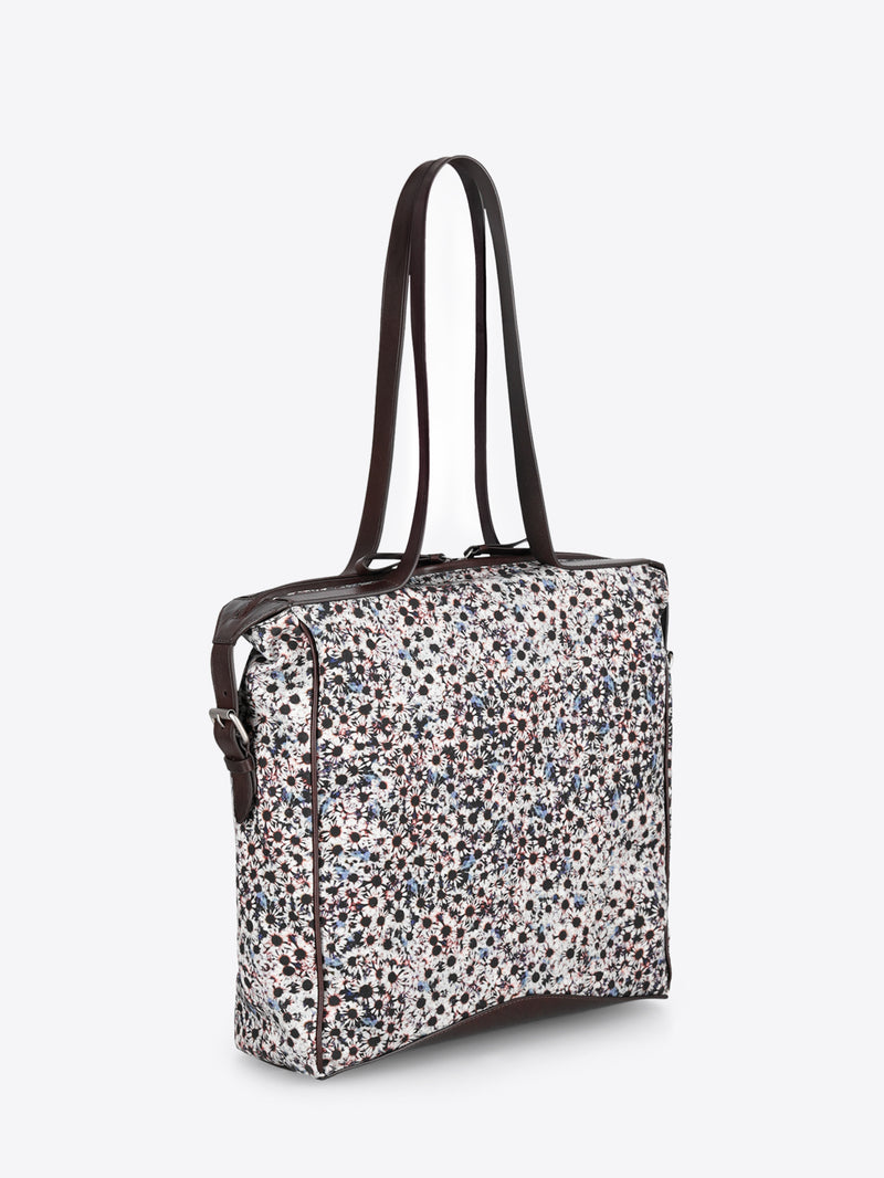 A structured tote bag with a dense multicolor floral print pattern on a light background, featuring dark brown faux-leather trim and shoulder straps.
