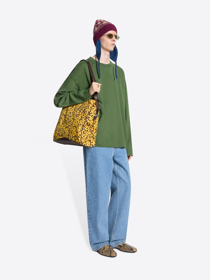 Person wearing a green sweater, blue jeans, and a colorful beanie holding a patterned yellow bag on a light gray background