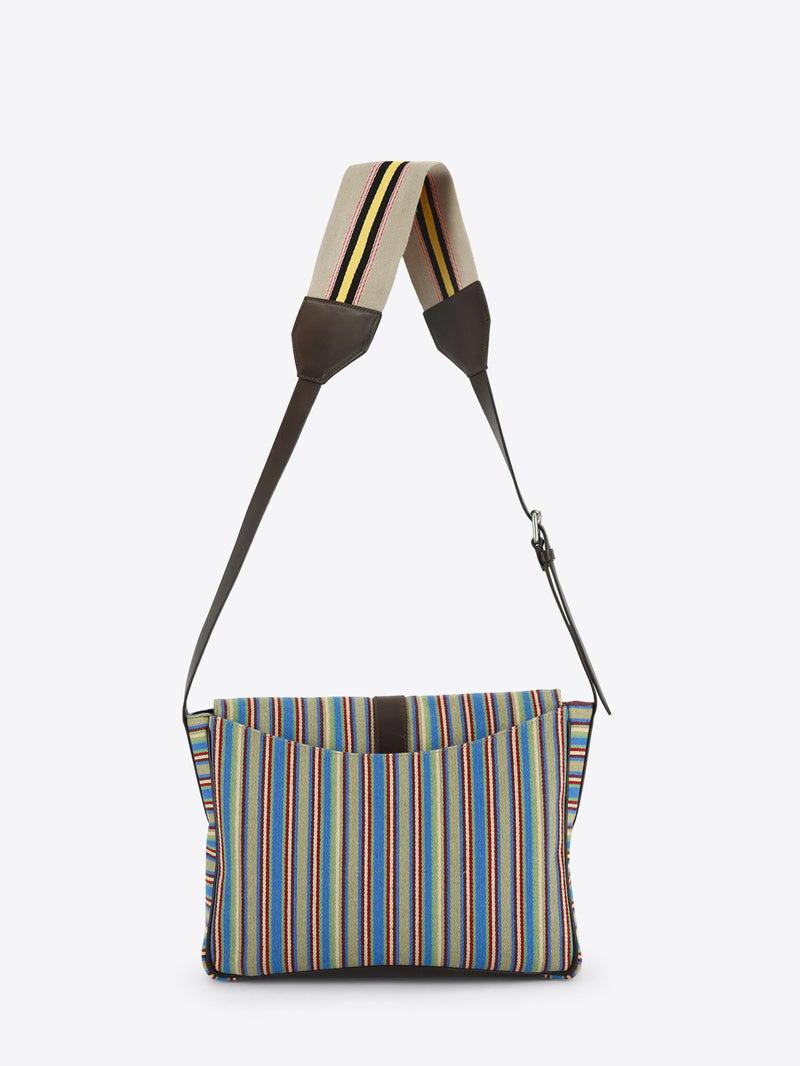 Back view of a multicolored striped shoulder bag with dark brown leather trim, a front buckle closure and an adjustable striped strap, shown against a light grey background.

